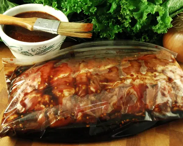 Karkówka w torebce, gotowa na grilla. W miseczce czeka aromatyczna marynata do karkówki na grilla, a obok pęczek zielonej sałaty i cebula.