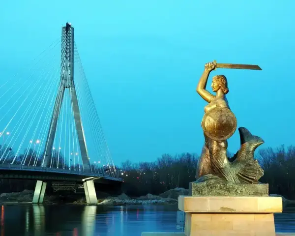Syrenka warszawska z mieczem i tarczą stoi nad Wisłą, w tle nowoczesny most.