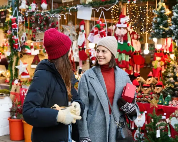 Två kvinnor njuter av julmarknader i Skåne, omgivna av julpynt och ljus. De håller presenter och ler mot varandra.