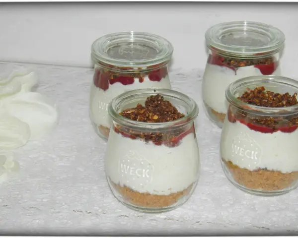 Vier leckere Desserts im Glas mit Krümelboden, cremiger Füllung, roter Fruchtsauce und knusprigem Topping.