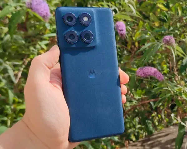 En hand håller en mörkblå motorola edge 60 fusion med fyra kameror mot en bakgrund av gröna blad och lila blommor.