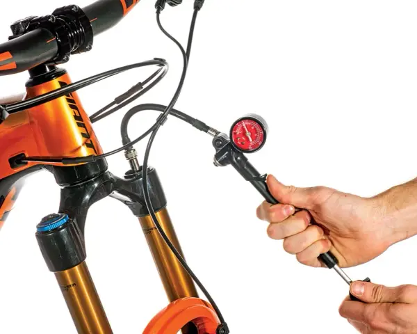 Adjusting the mtb suspension setup. A hand uses a shock pump with a gauge to set the air pressure on an orange mountain bike's front fork.