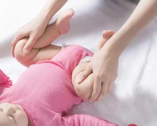 Dłonie delikatnie masują nóżki dziecka w różowym body, przygotowując do szerokiego pieluchowania.