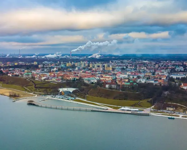Widok na Płock z lotu ptaka, z Wisłą w tle. Widać panoramę miasta, fabryki z dymiącymi kominami i nowoczesną infrastrukturę nad rzeką, która może być częścią atrakcji dla dzieci w Płocku.