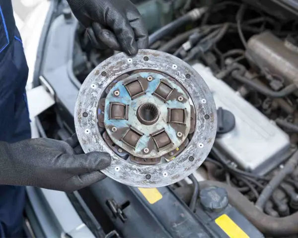 Mechanik w czarnych rękawiczkach trzyma zużyte sprzęgło samochodowe na tle silnika.
