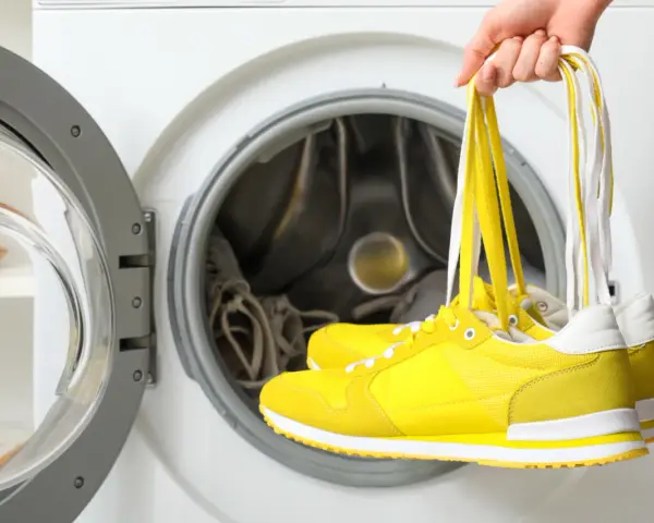 Jak prać buty? Ręka trzyma żółte trampki za sznurówki, gotowe do wrzucenia do otwartej pralki.