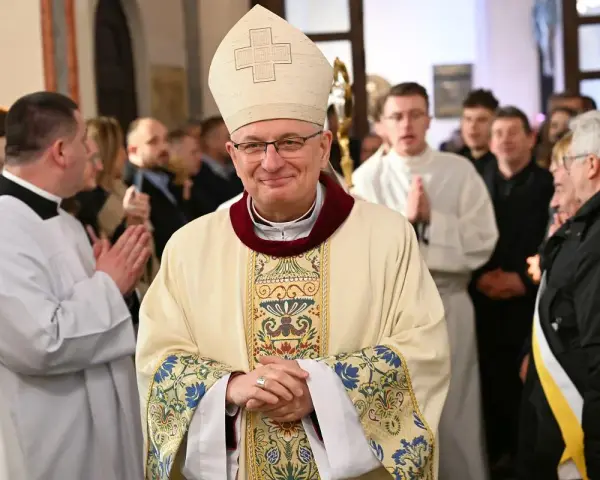 Uśmiechnięty biskup w stroju pontyfikalnym, otoczony przez wiernych i duchownych. W tle widać fragmenty kościoła, a na pierwszym planie szczegóły szat.