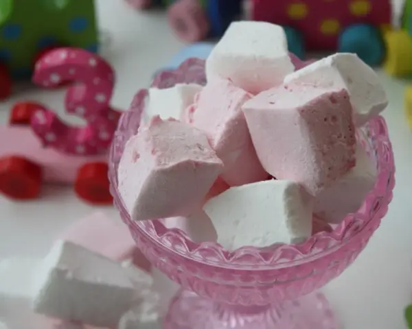En skål med hemmagjorda marshmallows i rosa och vitt. I bakgrunden syns färgglada träleksaker, som en rosa bil med siffran 3.