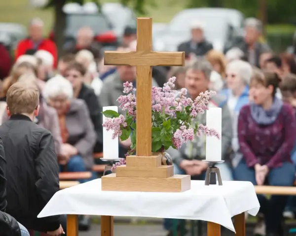 Holzkreuz mit Blumen und Kerzen vor einer Gemeinde. Ein Thema, das auch bei unterschiedlichen Konfessionen von Ehepartnern relevant ist: die Kirchensteuer.