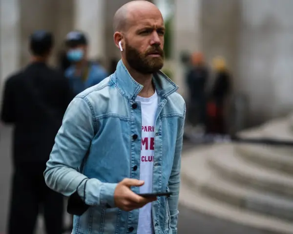 Hombre calvo sin barba, con auriculares y chaqueta vaquera, mira su móvil.