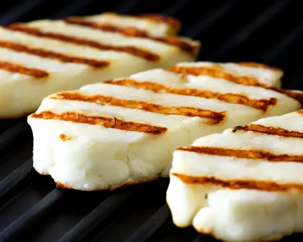 Trzy kawałki halloumi z grilla, z charakterystycznymi brązowymi paskami, leżą na czarnej, grillowej powierzchni.