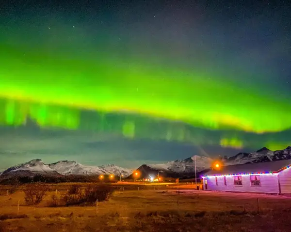 Islandia pogoda listopad: zorza polarna nad ośnieżonymi górami i domkiem z lampkami.
