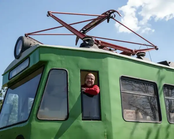 Uśmiechnięty mężczyzna w czerwonym swetrze wygląda z okna zielonej lokomotywy. To łódź Skierniewice, gotowa do podróży.
