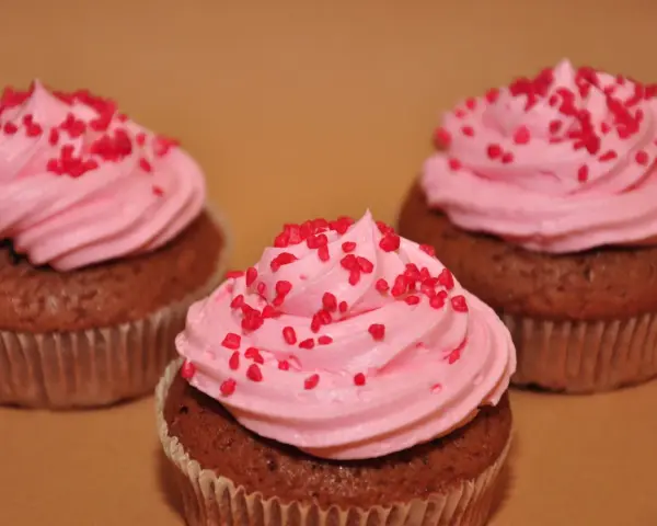 Tre chokladmuffins med rosa frosting och röda strössel, bakade i rosa muffinsformar.