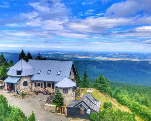 Kamienny schronisko z szarym dachem w Górach Izerskich, otoczone zielonymi lasami i rozległą panoramą dolin pod błękitnym, chmurowym niebem.