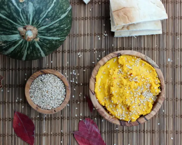 Ein leuchtend gelber Hummus, bestreut mit Sesam, wartet neben einer Schale voller Sesam und einem Kürbis. Perfekt für ein kleines Hummus-Baby-Festmahl!