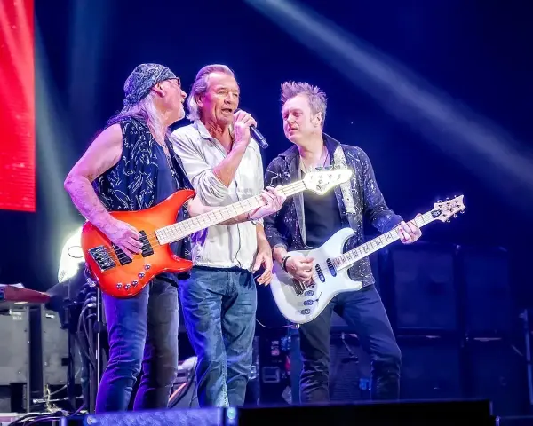 Trzech muzyków na scenie w głębokim fioletowym świetle Atlas Areny. Jeden śpiewa, dwaj grają na gitarach.