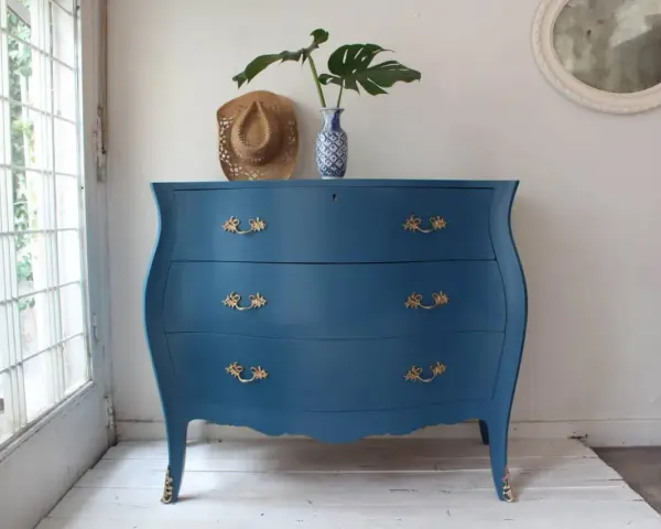 Cómoda antigua con **muebles pintados en azul** y tiradores dorados, decorada con un sombrero y un jarrón con hojas.