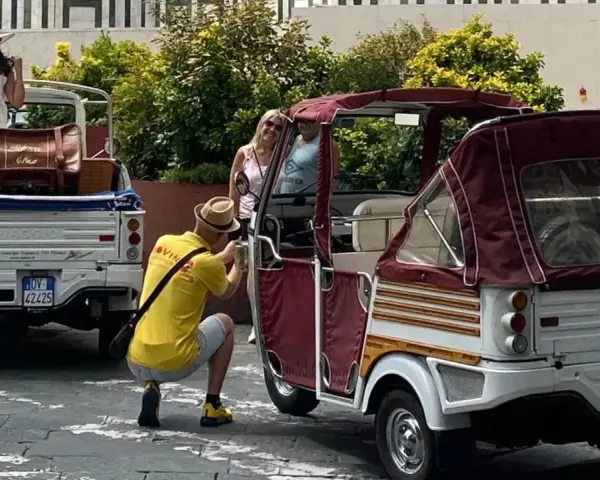 Turisti scattano foto a veicoli atipici, un Piaggio Ape e un tuk-tuk, in una strada soleggiata.