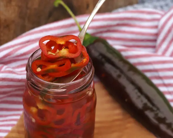 En burk med picklad chili, med en sked som lyfter upp en skiva. En mörk chili ligger bredvid.