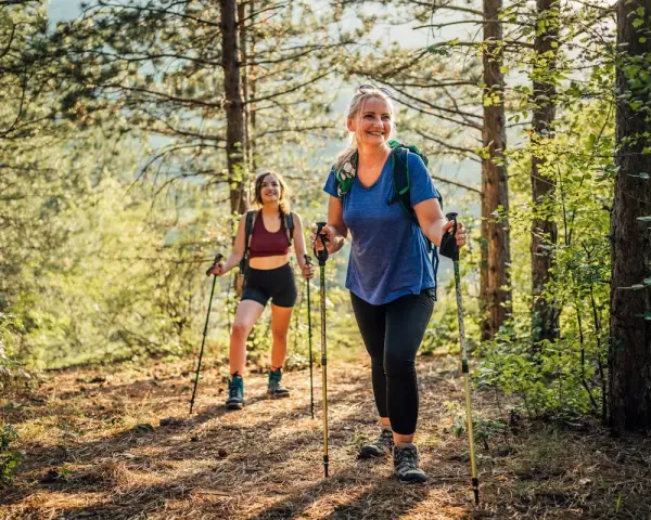 Deux femmes souriantes randonnent en forêt, utilisant des bâtons. Elles semblent apprécier la nature, un argument pour ou contre les bâtons de randonnée.