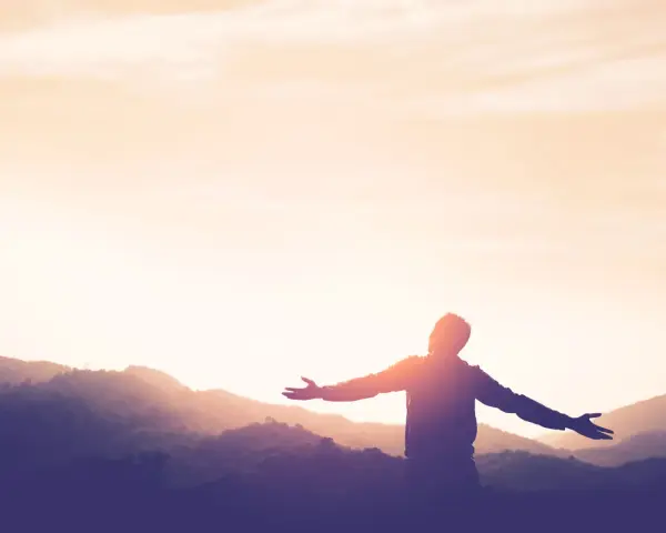 Silhouette einer Person mit ausgebreiteten Armen vor einer Bergkulisse bei Sonnenuntergang. Ein Symbol für Kraft und Hoffnung nach einer Krankheit.