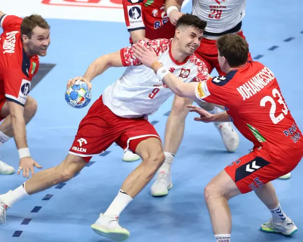 Gracz w białej koszulce z numerem 9 próbuje rzucić piłkę, podczas gdy obrońca w czerwonej koszulce z numerem 23 stara się mu przeszkodzić. Emocjonująca akcja w meczu piłki ręcznej.