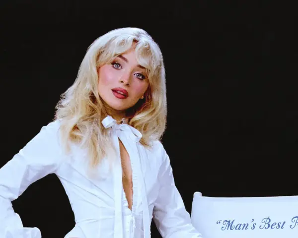 Sabrina Carpenter w białej koszuli, z rozpuszczonymi blond włosami, wygląda jak "man's best friend" na tle czerni.