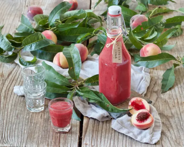 Flasche mit selbstgemachtem Pfirsichschnaps, frische Pfirsiche und Blätter auf Holztisch. Perfekt für den Sommer!