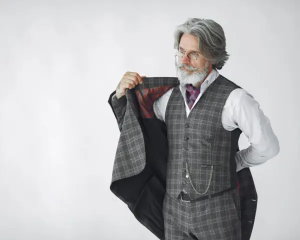 Elegancki mężczyzna z siwą brodą i wąsami, ubrany w kraciasty garnitur, prezentuje się jak prawdziwy arbiter elegantiarum.