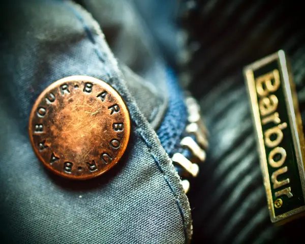 Detale kurtki Barbour: mosiężny guzik z logo i naszywka z nazwą firmy. Barbour to znana marka odzieży.
