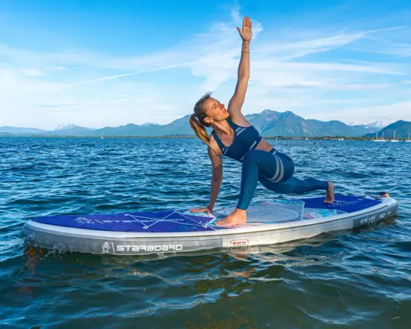 Frau praktiziert Krieger II auf einem SUP-Board auf ruhigem Wasser, Berge im Hintergrund.