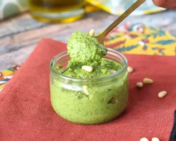 Łyżka nabiera aromatyczne pesto z rukoli z małego słoiczka. Na czerwonym tle widać orzeszki piniowe.