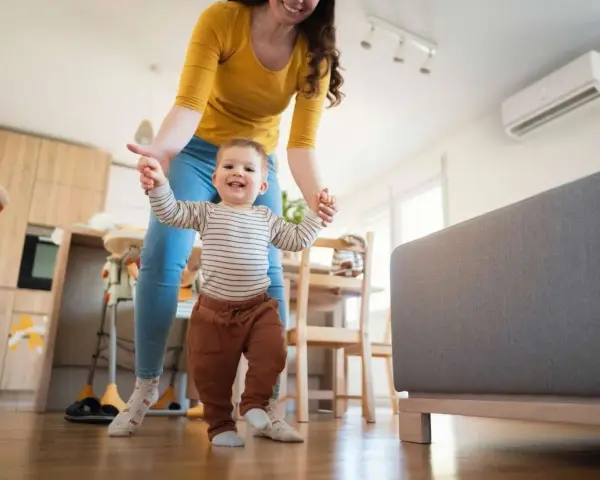 Mamá ayuda a su bebé a dar sus primeros pasos en una habitación cómoda, con el aire acondicionado bebes funcionando para mantener la temperatura ideal.
