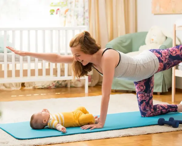 Mama wykonuje ćwiczenia po porodzie na macie, a jej maluch leży obok.