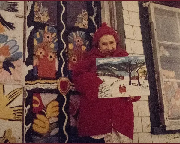 Maud Lewis, klädd i rött, håller upp en av sina charmiga vintermålningar framför sitt färgstarka hem.