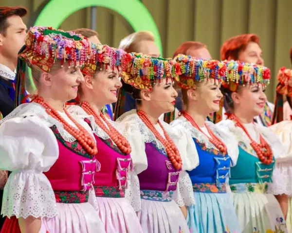 Szła dzieweczka w tradycyjnym stroju, z barwnym wiankiem na głowie, obok innych w ludowych strojach.