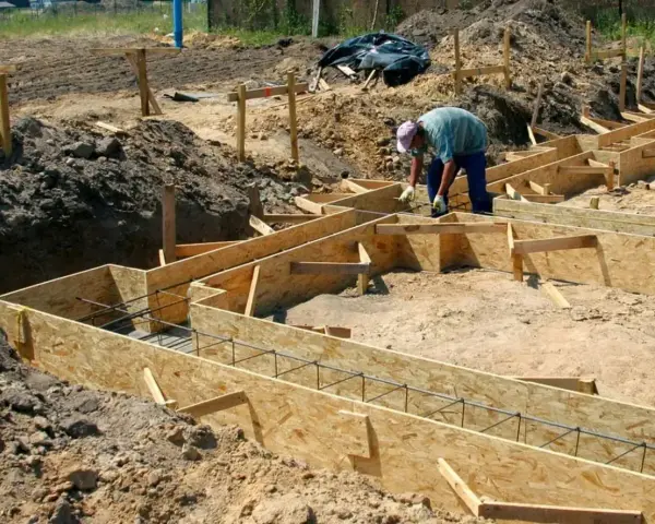 Budowa fundamentów: pracownik układa zbrojenie w drewnianym szalunku. Widać wiele desek na szalunek fundamentu, gotowych do zalania betonem.