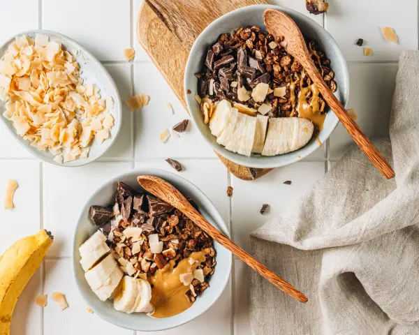 Due ciotole con granola fatta in casa, banana a fette, cioccolato fondente e burro d'arachidi. Cocco in scaglie a lato.