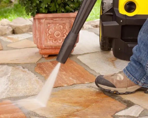 Pulire pavimento esterno con idropulitrice. Un getto d'acqua potente rimuove lo sporco dalle pietre del patio, mentre una persona in jeans e scarponi osserva.
