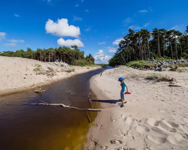 Chłopiec w kasku spaceruje po piaszczystym brzegu, gdzie wąski strumień wpada do wicie jezioro.