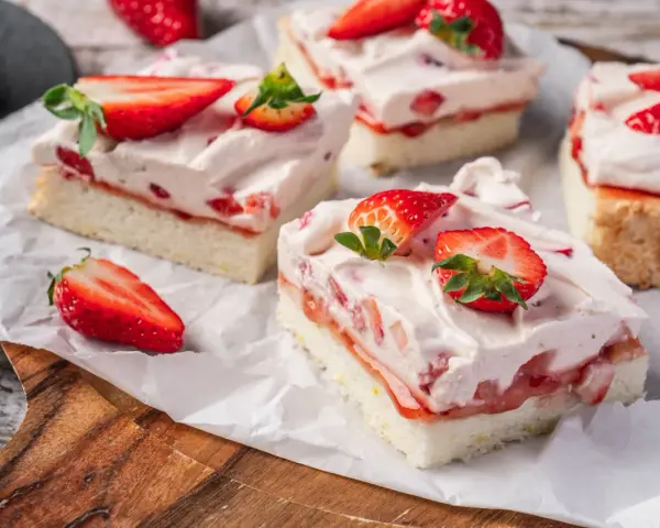 Saftige Stücke eines Erdbeerkuchen Blechs mit frischen Erdbeeren und Sahne, auf Backpapier angerichtet.