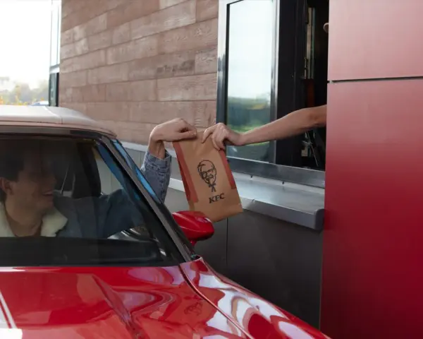 Mężczyzna w czerwonym aucie odbiera z okienka KFC torbę z jedzeniem. Wygląda na zadowolonego, że otrzymał swój mega pocket kcal.