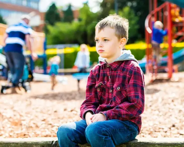 Chłopiec z zespołem Aspergera siedzi samotnie na placu zabaw, podczas gdy inne dzieci się bawią.