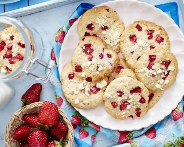 Saftige Erdbeer-Cookies mit weißen Schokostückchen auf einem Teller, daneben frische Erdbeeren und ein Glas Milch.