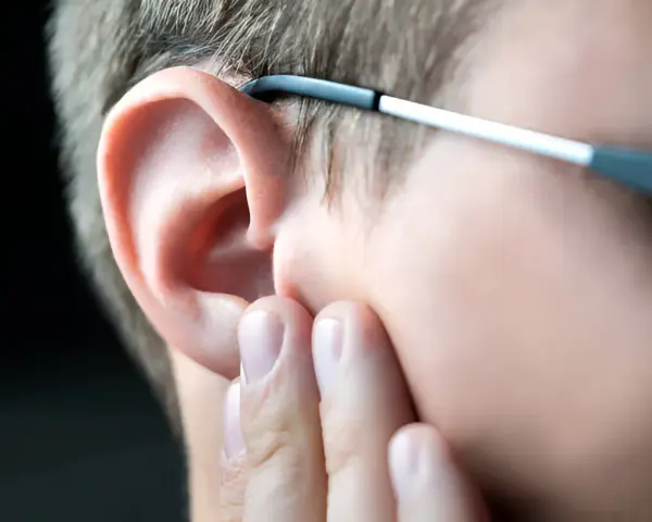 Dłoń dotyka ucha, sugerując uczucie zatkanego ucha. Okulary opierają się o skroń.