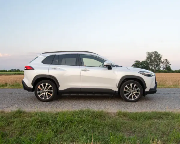 Biała Toyota Corolla Cross Hybrid stoi na poboczu drogi, otoczona polami i zieloną trawą.