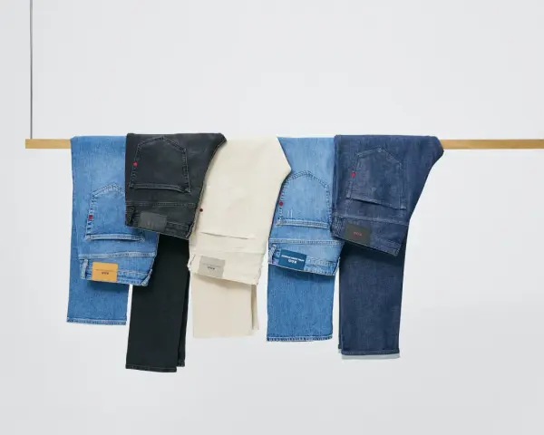 Cinco pares de pantalones vaqueros de distintos tonos cuelgan de una barra de madera. El **denim material** se presenta en azul claro, negro, beige, azul medio y azul oscuro.