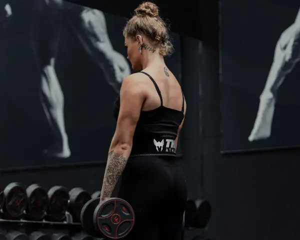 Kvinna med tatueringar utför shrugs med hantlar i ett gym.