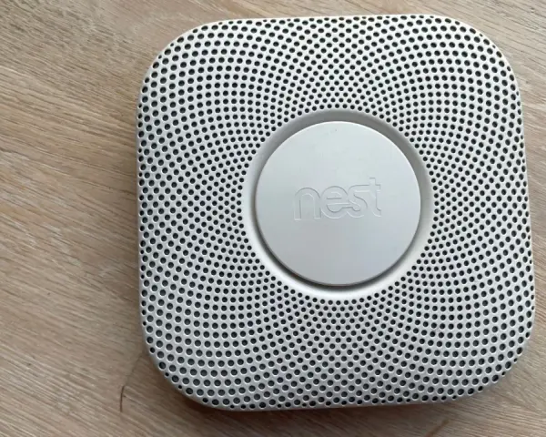 En vit Google Nest Protect-rökdetektor med ett runt "nest"-märke i mitten, placerad på ett trägolv.